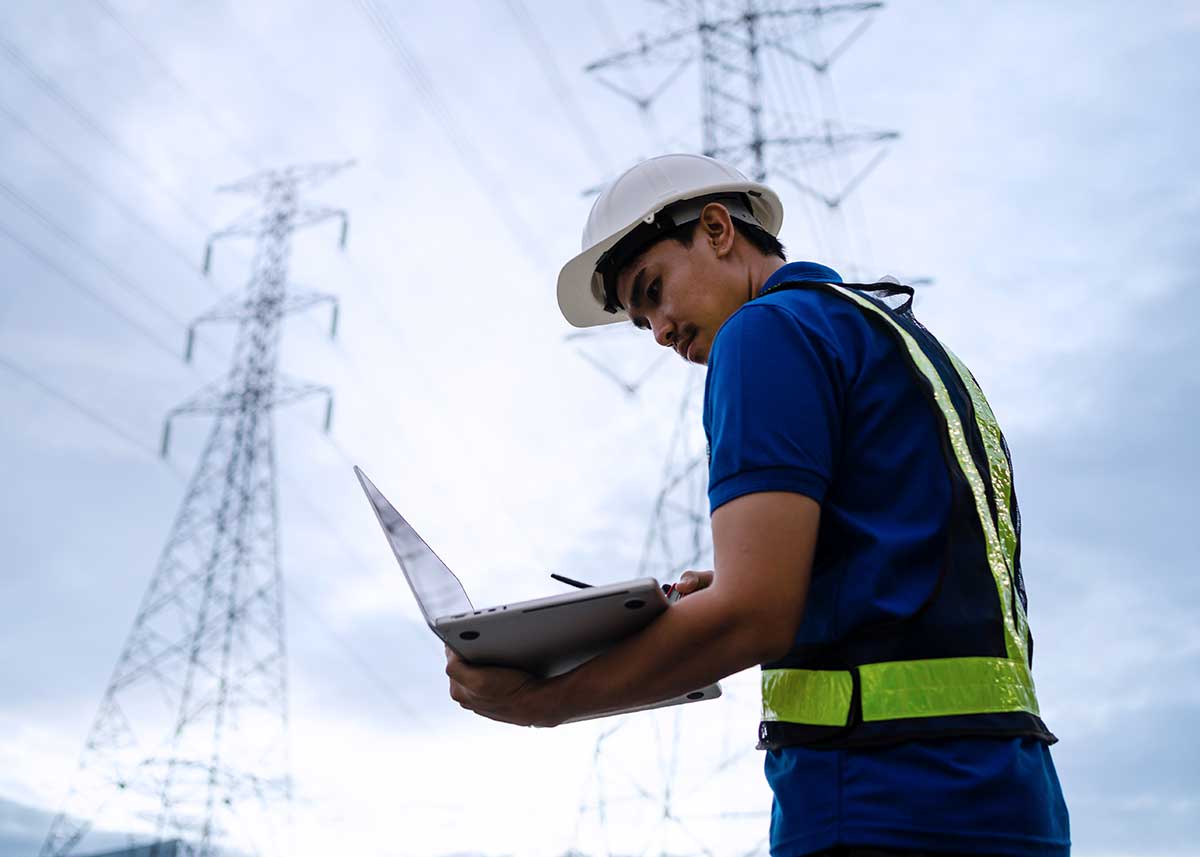 A man wearing a hard hat and safety vest is looking at a laptop while standing near a power line