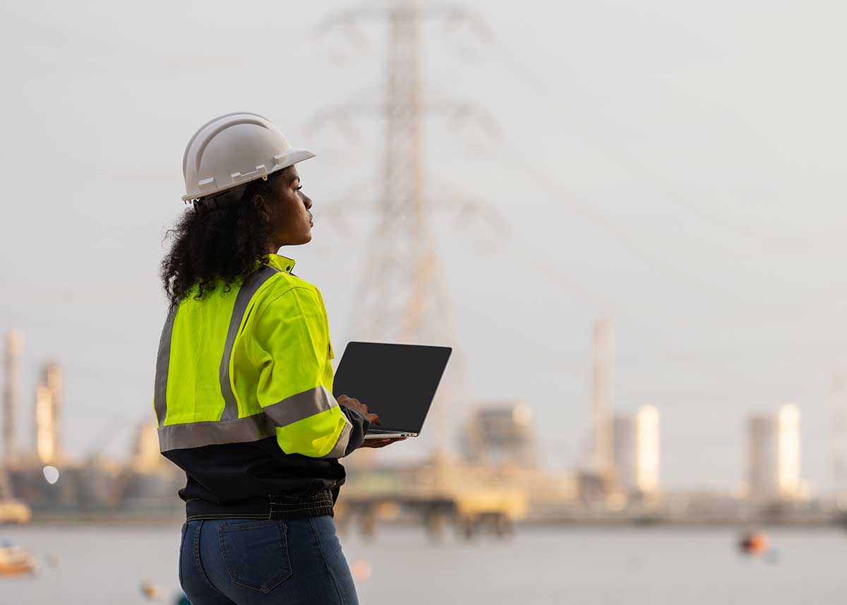 A female engineer in a high-visibility jacket and helmet uses a laptop and smartphone near a power plant and transmission towers, illustrating modern technology, communication, and industrial work