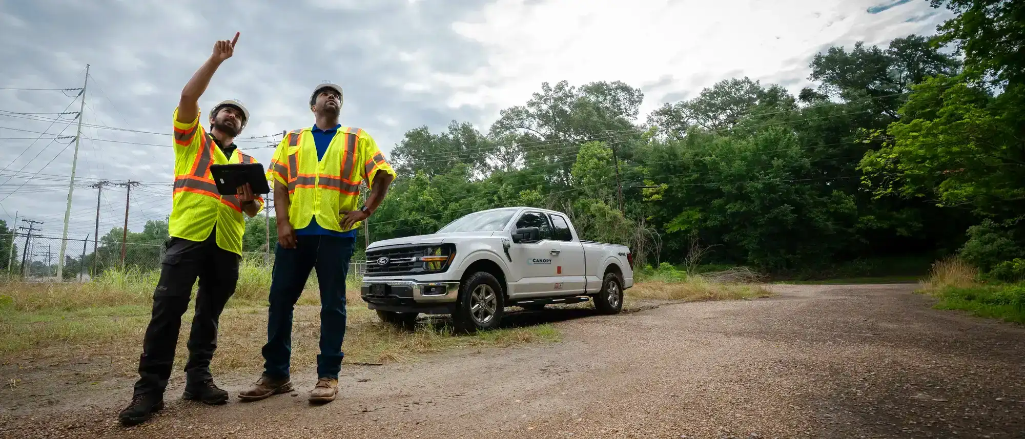 Two utility field professionals inspect overhead power lines near a wooded right-of-way, with one using a tablet.